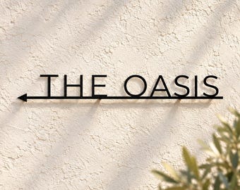 May include: A black metal wall sign featuring the text THE OASIS in a clean sans-serif font. The sign includes a horizontal line extending from the left side, ending in a directional arrow pointing to the left, mounted on a textured wall.