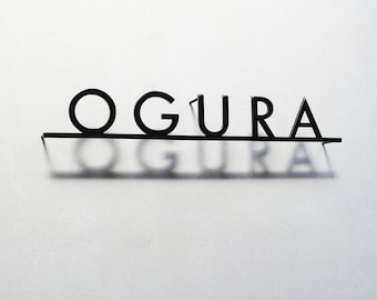 May include: Black metal sign spelling out the word "OGURA" in a sans-serif font. The letters are mounted on a thin horizontal bar, casting a shadow on the white wall behind it. The sign is a decorative element.