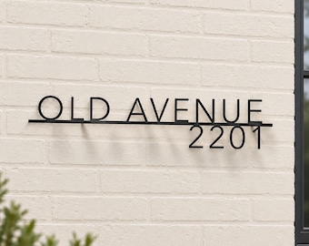 May include: A black metal wall sign featuring the text OLD AVENUE in capital letters positioned above a horizontal line, with the numbers 2201 displayed below the line on the right side, mounted on a light-colored brick wall surface.