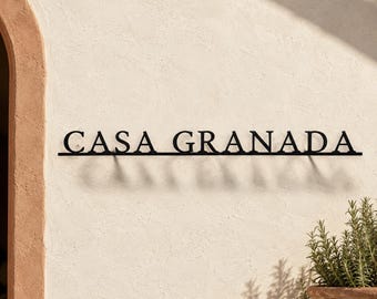 May include: A black metal wall sign featuring the text CASA GRANADA in a serif font. The letters are connected by a thin horizontal line at the base, creating a shadow effect against the light-colored wall surface behind the mounted signage.