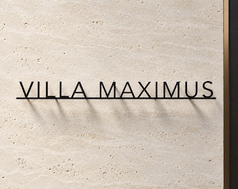 May include: A black metal wall sign featuring the text VILLA MAXIMUS in a clean, sans-serif font. The letters are connected by a thin horizontal line at the base, creating a floating effect against the textured, light beige stone wall surface.