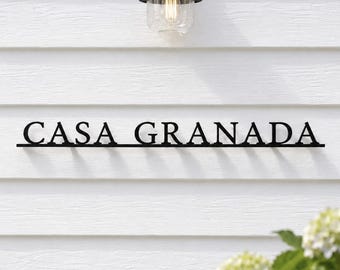 May include: A black metal wall sign featuring the text CASA GRANADA in a serif font, mounted on white horizontal siding. The sign is positioned below a black outdoor light fixture with a clear glass shade and a visible filament bulb.