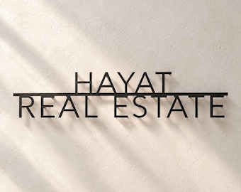 May include: A black metal wall sign featuring the text HAYAT REAL ESTATE in a clean, sans-serif font. The letters are mounted on a horizontal bar, creating a dimensional effect with shadows cast against the textured wall surface.