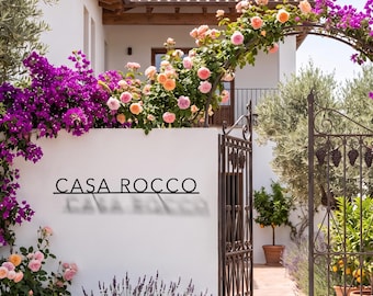 May include: A white stucco wall features the black metal text CASA ROCCO. An arched metal gate stands beside the wall, which is adorned with vibrant purple bougainvillea and soft peach roses. Lavender plants grow at the base of the wall.
