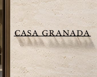 May include: A black metal wall sign featuring the text CASA GRANADA in a serif font. The sign is mounted on a light beige textured stone surface, creating a distinct shadow beneath the letters that emphasizes the depth of the metal lettering.