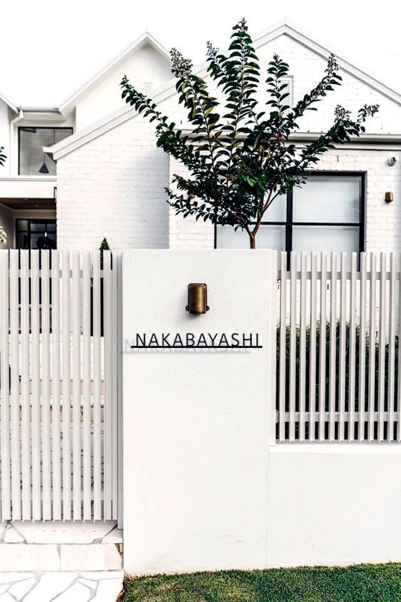 May include: A white picket fence with a gate and a white brick wall with a brass light fixture and a black sign that reads "NAKABAYASHI".