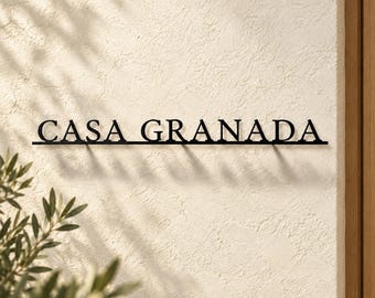May include: A black metal wall sign featuring the text CASA GRANADA in a serif font. The letters are connected by a thin horizontal line at the base, creating a clean and structured appearance against the textured off-white wall surface.