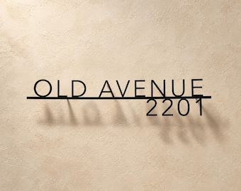 May include: A black metal wall sign featuring the text OLD AVENUE in capital letters on the top line and the number 2201 on the bottom line, with a horizontal bar separating the two lines of text and casting a shadow on the wall.