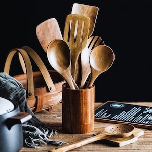 May include: A selection of wooden kitchen utensils, such as spoons and spatulas, presented in a wooden container. The utensils display a natural wood grain and are arranged on a wooden surface, evoking a rustic kitchen aesthetic.