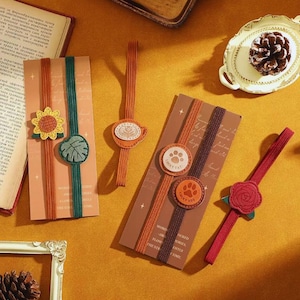 May include: A collection of decorative bookmarks with various designs. The bookmarks feature a sunflower, leaf, coffee cup, paw prints, and a rose. Each bookmark has a decorative element attached to a ribbon. The text "WORD FLOW STORIES" is visible on one bookmark.