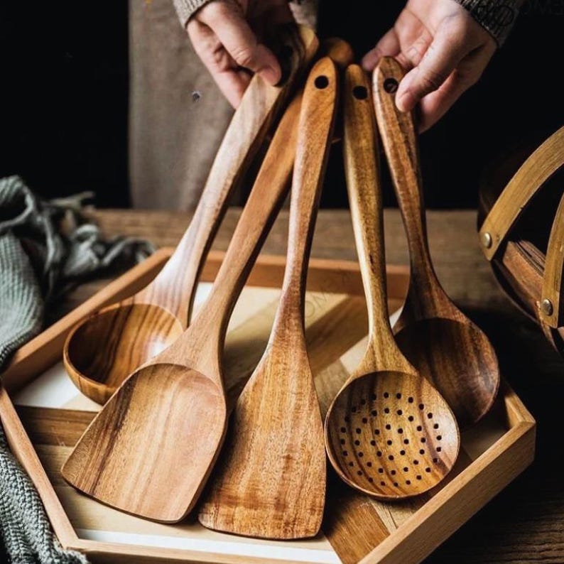 May include: A selection of wooden kitchen utensils, including a slotted spoon, spatula, and ladle, presented in a wooden tray. The utensils are crafted from light brown wood and feature long handles. The image is captured from a slightly elevated perspective.