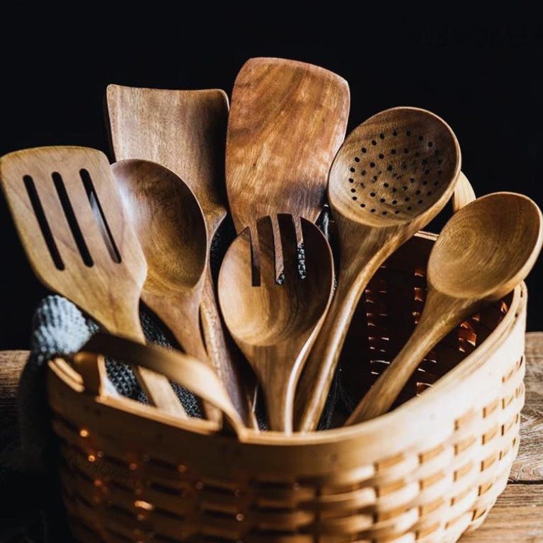 May include: A selection of wooden kitchen utensils, including spoons, spatulas, and slotted tools, are arranged in a woven basket. The utensils are made of light brown wood and have various shapes for cooking and serving.