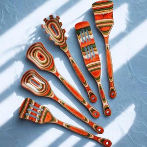 Op de afbeelding: Een set van zes houten keukengerei met een levendig, gelaagd ontwerp in tinten van oranje, rood, groen en bruin. De set bevat een spatel, een sleuflepel, een vork en ander kookgerei. De handvatten zijn effen oranje.