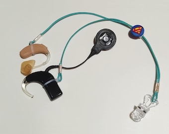 May include: A collection of hearing aids and related accessories. The image shows a beige and black hearing aid, a black receiver, and a clear clip. A teal cord connects the components, with a Superman logo charm.