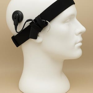 May include: A black headband with attached earphone and a small black device, worn on a white mannequin head. The device has a curved metal piece that fits over the ear. The background is a solid tan color.