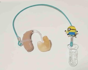 May include: A beige hearing aid with a clear plastic tube and ear mold, connected to a teal cord. A yellow Minion character charm is attached to the cord, which ends in a clear plastic clip.