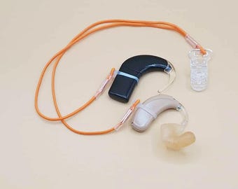 May include: Two hearing aids, one black and one beige, with clear tubing and ear molds. An orange lanyard connects the aids to a clear plastic clip. The aids are designed to assist with hearing.