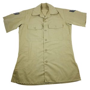 May include: Tan short-sleeved shirt with two chest pockets and a collared neckline. The shirt has a button-down front and epaulettes with black stripes on both sleeves. The shirt is made of a light-colored fabric.