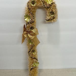 May include: A gold-colored, candy cane-shaped Christmas decoration. The decoration is covered in gold tinsel and adorned with gold-colored flowers, beads, and a bow. The candy cane is approximately 12 inches tall.