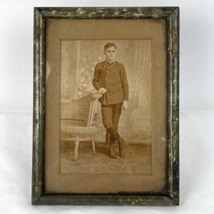 May include: A rectangular picture frame with a distressed dark metallic finish containing a sepia-toned vintage photograph. The image depicts a person standing beside a wicker chair, wearing a double-breasted jacket, breeches, and tall boots.