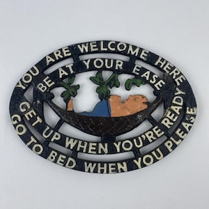 May include: Oval, cast iron sign with the words "YOU ARE WELCOME HERE BE AT YOUR EASE GET UP WHEN YOU'RE READY GO TO BED WHEN YOU PLEASE". The sign has a hammock with a person in it and green leaves.