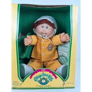 May include: A 1985 Cabbage Patch Kids doll in its original packaging. The doll has brown yarn hair, a white knit cap, and a yellow long-sleeved shirt with a small bear graphic and matching yellow pants. It wears white shoes and sits in a green box.