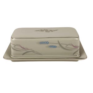 May include: A rectangular, off-white butter dish with a lid, featuring a silver trim and wheat-like design in blue, gray, and pink. The dish sits on a matching rectangular base, creating a cohesive set.