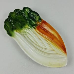 May include: A ceramic serving dish shaped like a bunch of bok choy with vibrant green leaves, a white stalk, and orange-tinted roots. The piece features a glossy finish with molded textures that mimic the natural appearance of the vegetable.