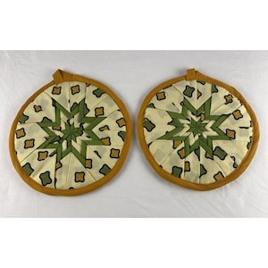 May include: Two round fabric pot holders featuring a folded star pattern in shades of olive green, mustard yellow, and cream. Each circular piece is finished with a solid mustard yellow fabric border and a small fabric loop for hanging.