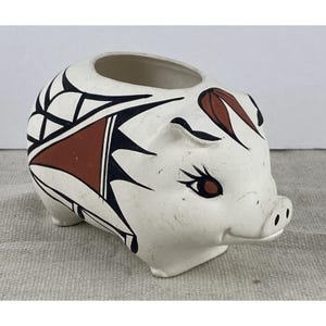 May include: A ceramic piggy bank shaped like a pig with a matte off-white finish. The body features hand-painted geometric patterns in black and terracotta red. The pig has stylized eyes with long lashes and a snout with two small circular nostrils.