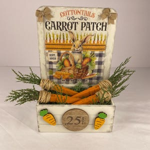 May include: A white wooden box with orange carrot decorations and a sign that reads "Carrot Patch." The sign features a rabbit with a basket of carrots. The box contains faux carrots and greenery, perfect for Easter decor.