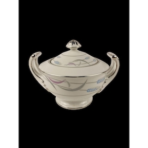 May include: A white ceramic sugar bowl with a lid, featuring silver trim and handles. The bowl is decorated with a pattern of blue, lavender, and gray wheat stalks. The lid has a small silver knob.