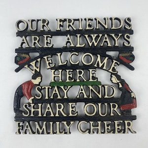 May include: Black cast iron sign with white lettering that reads "OUR FRIENDS ARE ALWAYS WELCOME HERE STAY AND SHARE OUR FAMILY CHEER." The sign has a decorative design with figures and accents of red, green, and black.