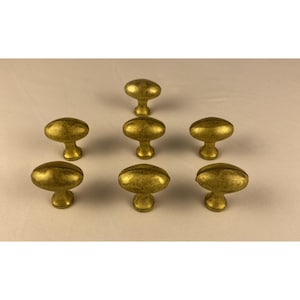May include: A collection of seven oval-shaped cabinet knobs made of brass with a distressed, antique gold finish. Each knob features a smooth, rounded top surface and a tapered base, arranged in a symmetrical pattern on a neutral surface.