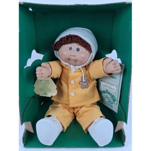 May include: A Cabbage Patch Kids doll with brown yarn hair and a white knit cap, wearing a yellow long-sleeved shirt with three white buttons and a small bear graphic, matching yellow pants, and white shoes, presented inside a green cardboard box.