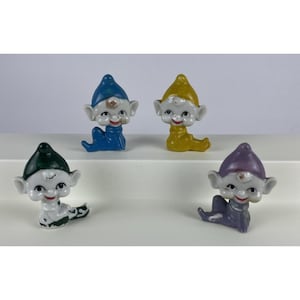 May include: Four small ceramic elf figurines in a seated position. Each elf has a white face, large ears, and a pointed hat. The hats and clothing are colored in blue, yellow, dark green, and purple. Each figure features painted facial details.