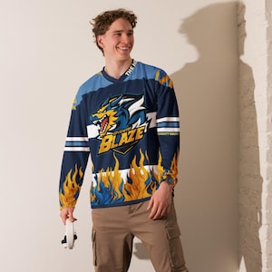 Puede incluir: Una camiseta de manga larga azul y amarilla con un gráfico de lobo y el texto "Coventry Blaze" en la parte delantera. La camiseta tiene llamas en las mangas y el texto "The Blaze" en el cuello.