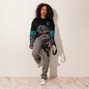 May include: A black and teal hockey jersey with a graphic of a hockey player and the text "Belfast Giants". The jersey has a white C on the chest and a white stripe on each sleeve.