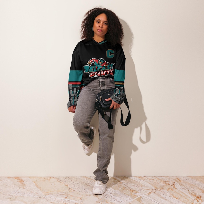 May include: A black and teal hockey jersey with a graphic of a hockey player and the text "Belfast Giants". The jersey has a white C on the chest and a white stripe on each sleeve.