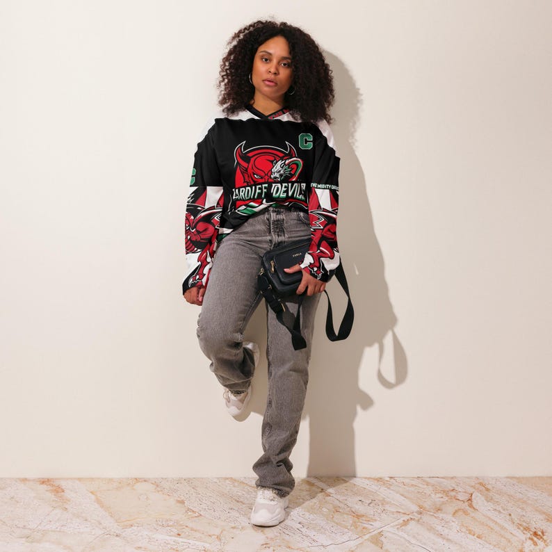 May include: A woman wearing a black, white, and red long-sleeved jersey with a graphic of a red devil and the text "CARDIFF DEVILS". She is also wearing gray jeans and a black crossbody bag.
