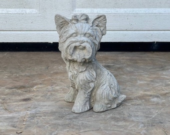 May include: A gray concrete statue of a Yorkshire Terrier dog sitting with its head tilted to the side. The dog has a bow on its head and is detailed with fur.