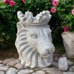 May include: A grey stone sculpture of a lion head wearing a crown. The lion features detailed carved fur, pointed ears, and a textured mane. The crown sits atop the head with distinct scalloped edges and a beaded band detail around the base.