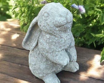 May include: A gray stone rabbit statue with long ears and a smiling face sits on a wooden surface. The bunny is in a seated position with its paws together. Green foliage and purple flowers are in the background, creating a garden theme.