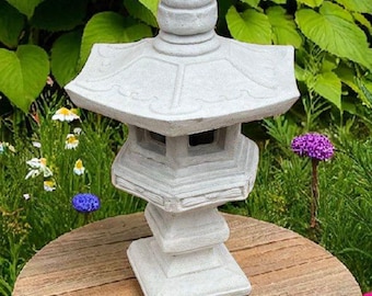 Può includere: Una lanterna da giardino grigia in stile pagoda è posizionata su un tavolo rotondo in legno, circondata da fiori colorati e vegetazione. La lanterna ha un design a più livelli con una parte superiore a cupola.