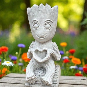May include: A gray statue of a character with a smiling face and a heart-shaped design on its chest. The statue is sitting on a wooden bench surrounded by colorful flowers. The background is a blurred view of a garden.