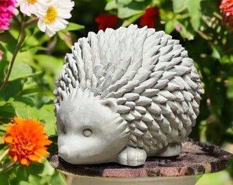 May include: A gray concrete hedgehog statue with a detailed spiky design sits on a gray pedestal in a garden setting. The statue is surrounded by pink, white, and orange flowers.