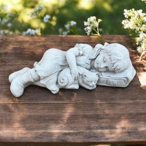 May include: A light-coloured sculpture of a sleeping child and a cat. The child lies on a pillow, with the cat curled up beside them. The sculpture is placed on a wooden table, with a blurred green background.