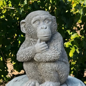 May include: A gray concrete statue of a small monkey sitting with its arms crossed. The monkey is sitting on a round, gray concrete base.