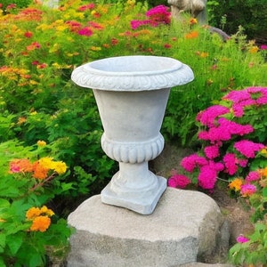 Peut inclure: Une urne de jardin en pierre grise avec un bord décoratif est posée sur un gros rocher dans un jardin. L'urne est entourée de fleurs colorées.