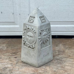 May include: A gray concrete obelisk with a sun face on each side. The obelisk is labeled "South" and "East" and has the letters "S" and "E" on the top.